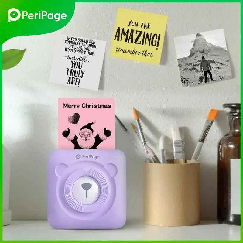 PeriPage A6 Mini Printer in purple bear design printing a Merry Christmas note, placed on a desk with brushes and sticky notes on the wall.